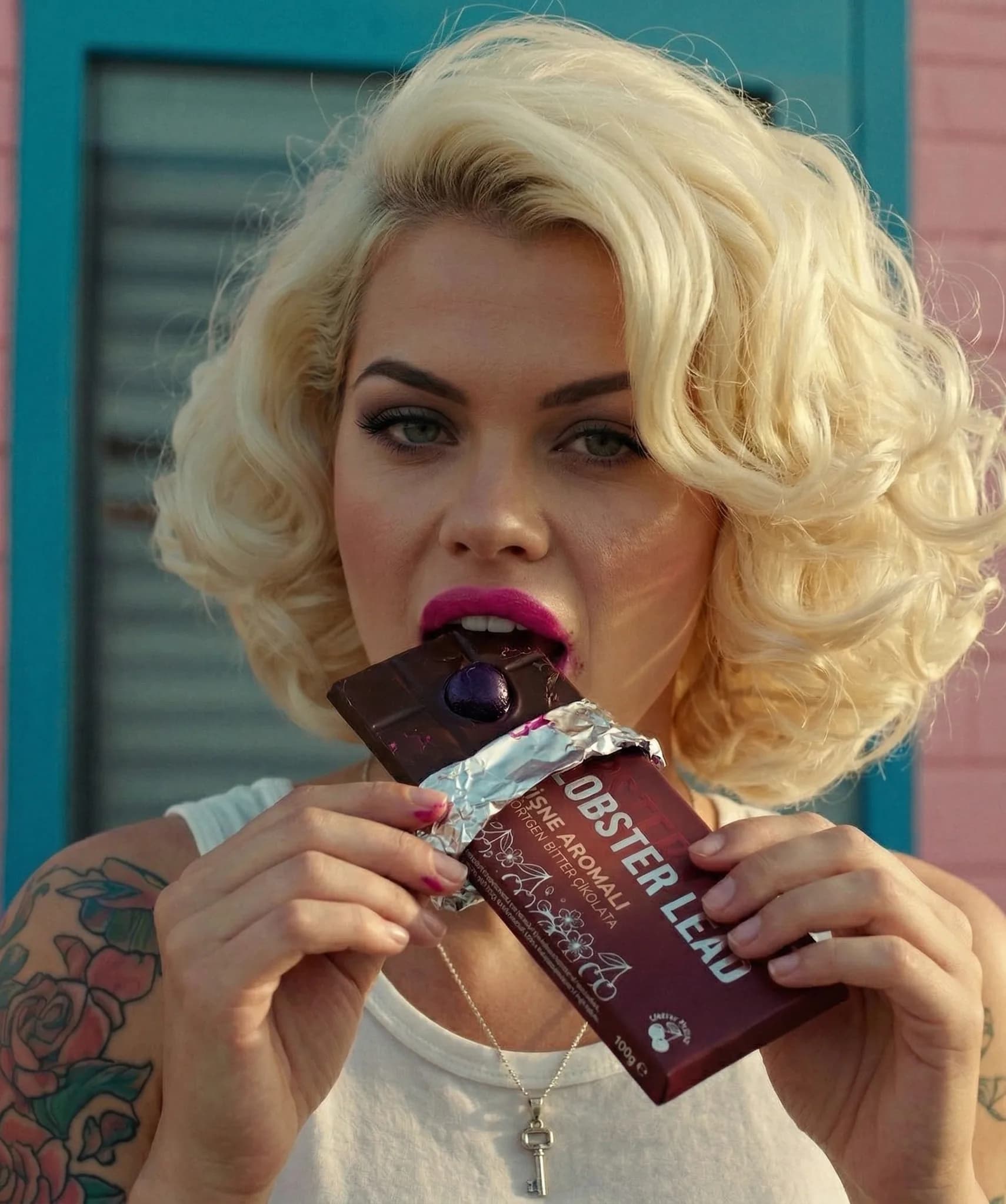A woman with retro hair and tattoos bites a Lobster Lead chocolate bar in front of a coloured wall